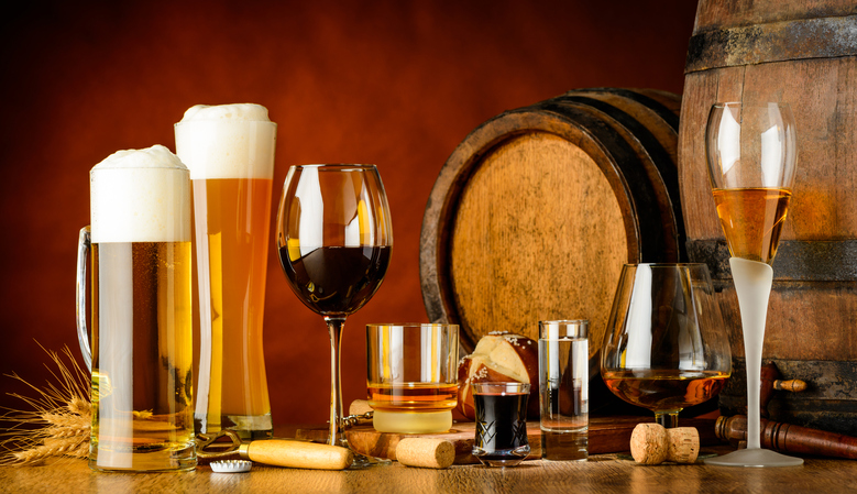 alcoholic drinks on wooden table in glasses, mugs and shots with barrel in background